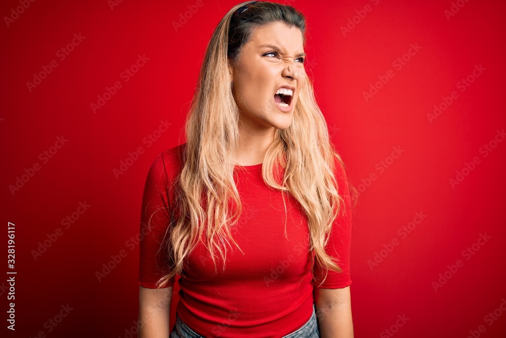 Young beautiful blonde woman wearing casual t-shirt standing over isolated red background angry and mad screaming frustrated and furious, shouting with anger. Rage and aggressive concept. Young beautiful blonde woman wearing casual t-shirt standing over isolated red background angry and mad screaming frustrated and furious, shouting with anger. Rage and aggressive concept.