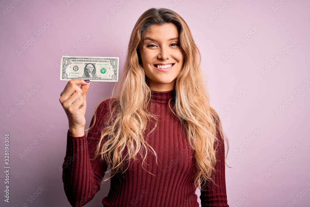 Young beautiful blonde woman holding one dollar banknote over isolated pink background with a happy face standing and smiling with a confident smile showing teeth Young beautiful blonde woman holding one dollar banknote over isolated pink background with a happy face standing and smiling with a confident smile showing teeth