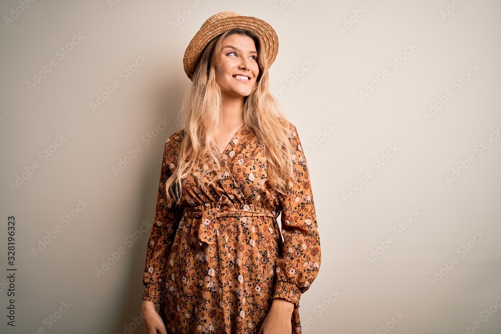 Young beautiful blonde woman wearing summer dress and hat over isolated white background looking away to side with smile on face, natural expression. Laughing confident. Young beautiful blonde woman wearing summer dress and hat over isolated white background looking away to side with smile on face, natural expression. Laughing confident.