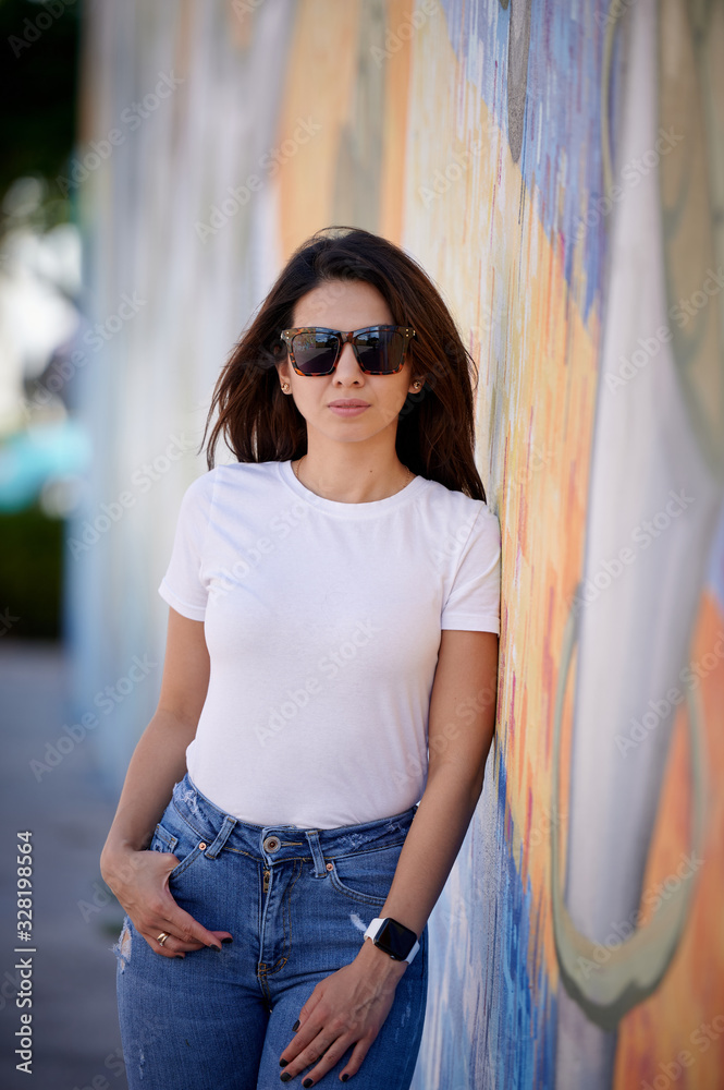 Smiling woman in sunglasses with the right hand in the pocket standing on the street next to the wall