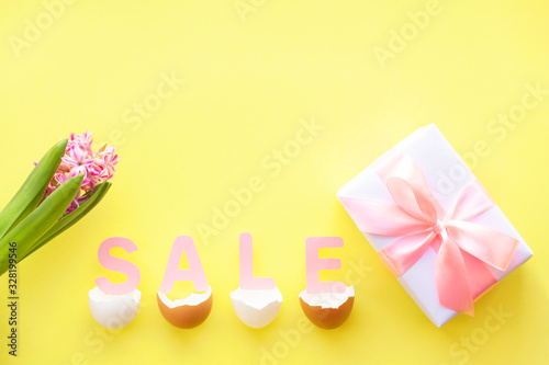 Pink letters SALE lie in broken egg shell, hyacinth flower and white gift box on yellow background. Spring, Easter sale concept. Flat lay, top view, copy space.