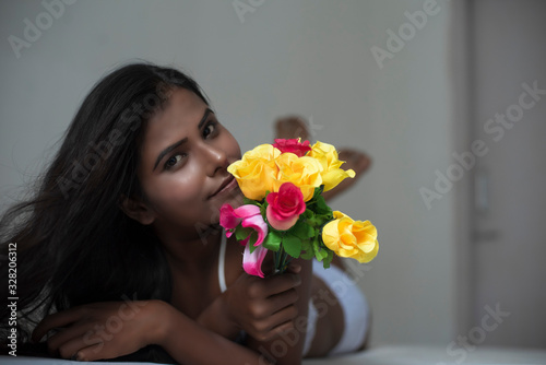 Portrait of an young dark skinned Indian Bengali woman in lingerie and vibrant flowers lying on  white bed in casual mood in white background. Indian lifestyle and  boudoir photography.