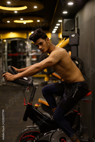 An young and handsome Indian Bengali brunette man with muscular body doing cycling in a multi gym. Fitness and Indian lifestyle.