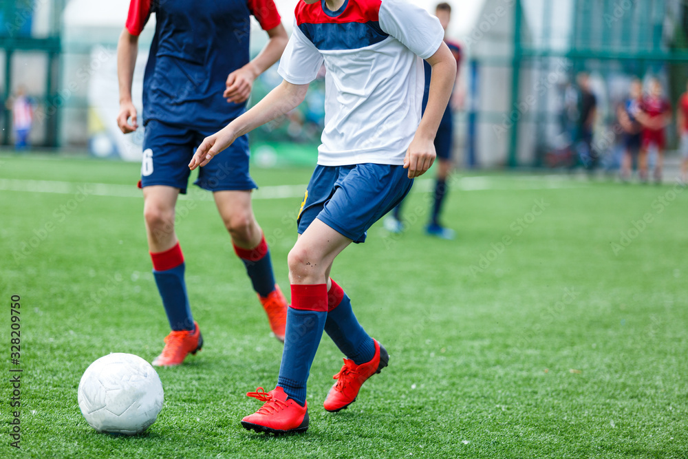 Obraz premium Boys in white and blue sportswear plays football on field, dribbles ball. Young soccer players with ball on green grass. Training, football, active lifestyle for kids concept 