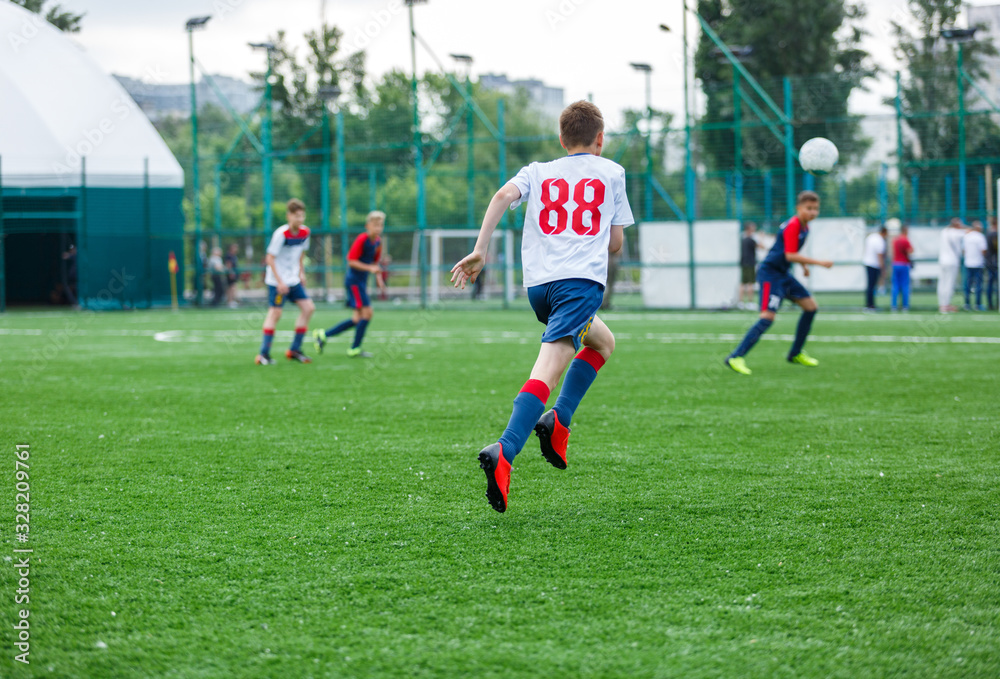 Obraz premium Boys in white and blue sportswear plays football on field, dribbles ball. Young soccer players with ball on green grass. Training, football, active lifestyle for kids concept