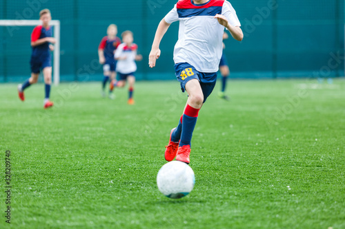 Boys in white and blue sportswear plays football on field, dribbles ball. Young soccer players with ball on green grass. Training, football, active lifestyle for kids concept 