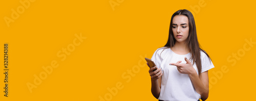 Thoughtful caucasian lady is pointing to her phone while posing on a yellow background with blank space © Strelciuc