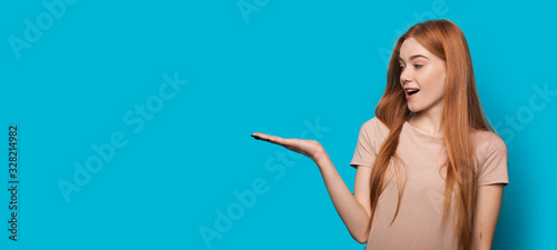 Ginger caucasian lady with freckles is holding something on her palm while posing on a blue wall with blank space