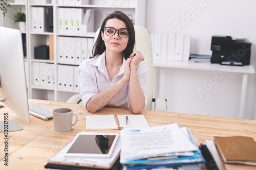 Portrait of beautiful female manager in glasses sitting at wooden desk with open diary in modern office