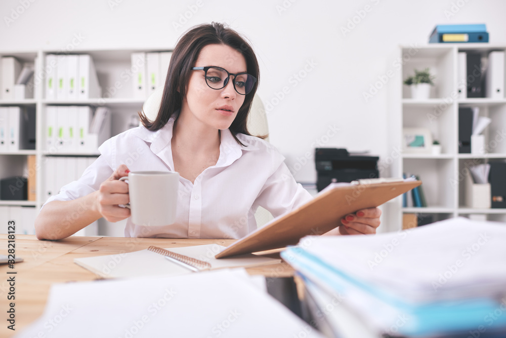 Busy young office woman sitting at table and reading papers while drinking coffee in office Busy young office woman sitting at table and reading papers while drinking coffee in office