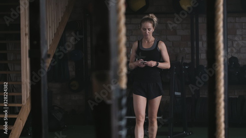 Young beautiful athletic blonde woman walking along large empty gym with smartphone preparing to exercise slow motion.