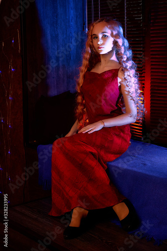 Portrait of young elegant tender blonde teenage girl in red dress on the window sill in dark room with blue light. Caucasian female model with very long hair posing indoors