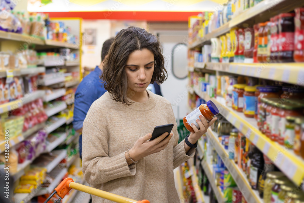 Focused woman using smartphone in grocery store. Young woman reading checklist via smartphone and choosing products in grocery store. Supermarket concept