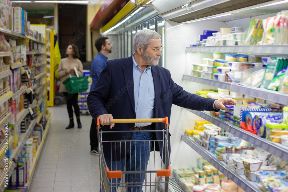 Mature man choosing dairy products in supermarket. Serious man with shopping trolley looking at shelves in refrigerator while buying goods in store. Shopping concept
