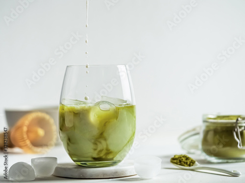 Iced Matcha Latte Tea with pouring milk drops into tumbler glass. Matcha latte and ingredients on white marble background. Copy space for txt or design