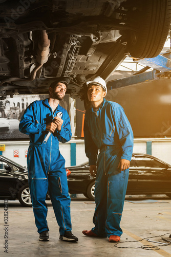 Mechanic in blue work wear uniform inspects the car bottom with a wrench with his assistant. Automobile repairing service, Professional occupation teamwork. Vehicle maintenance. Verticle image.
