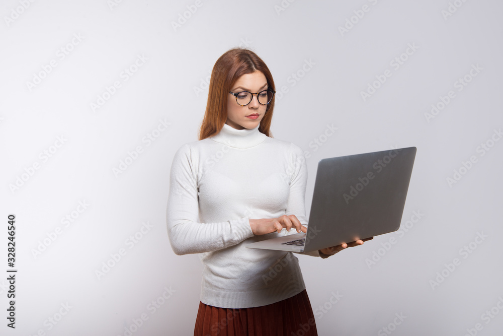 Fototapeta premium Serious young businesswoman using laptop. Focused young woman in eyeglasses standing and using laptop computer isolated on white background. Technology concept