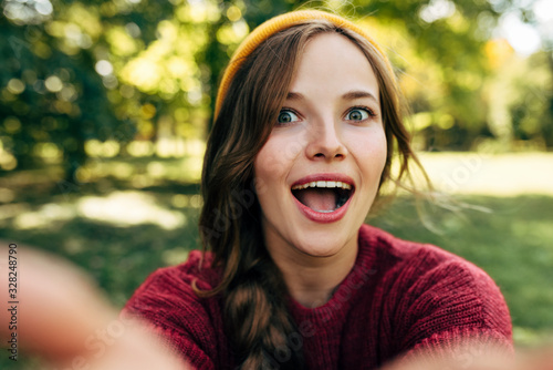 Close-up self portrait of happy young blonde woman smiling broadly, has surprised expression, posing on nature background in the park. Pretty female taking selfie. People, travel, lifestyle