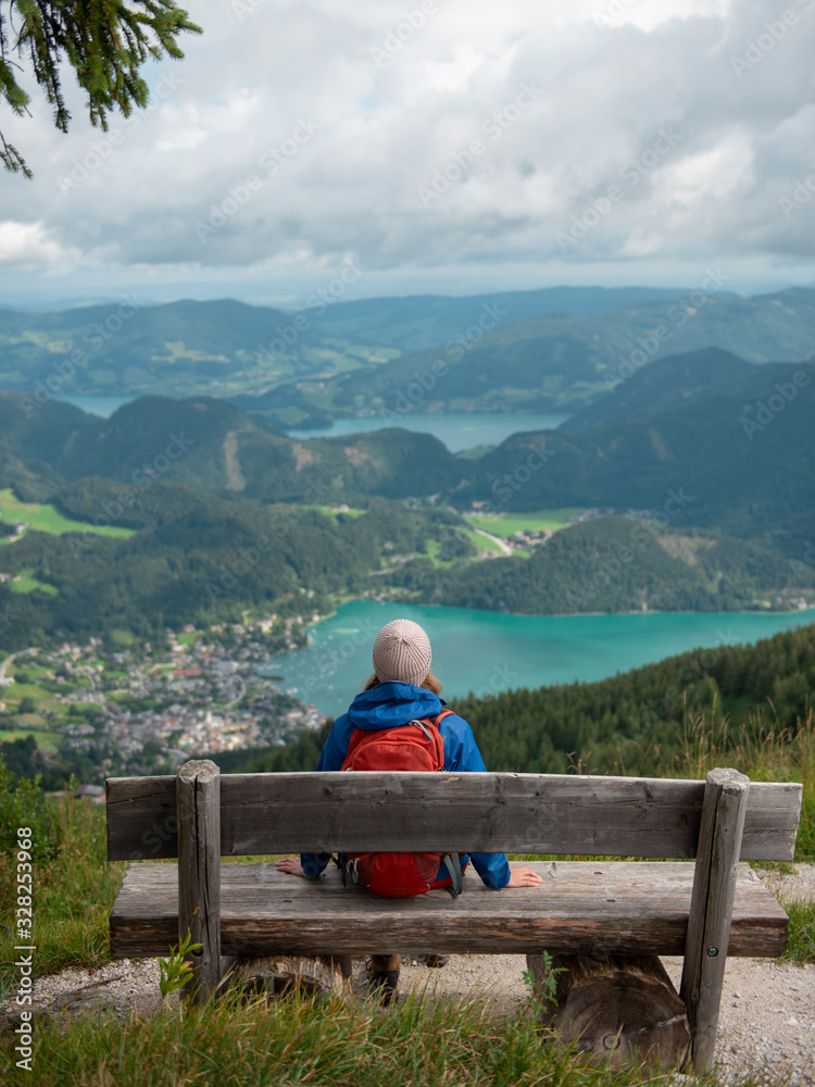 Woman with backpack sitting on wooden bench under tree in mountains enjoying scenic view