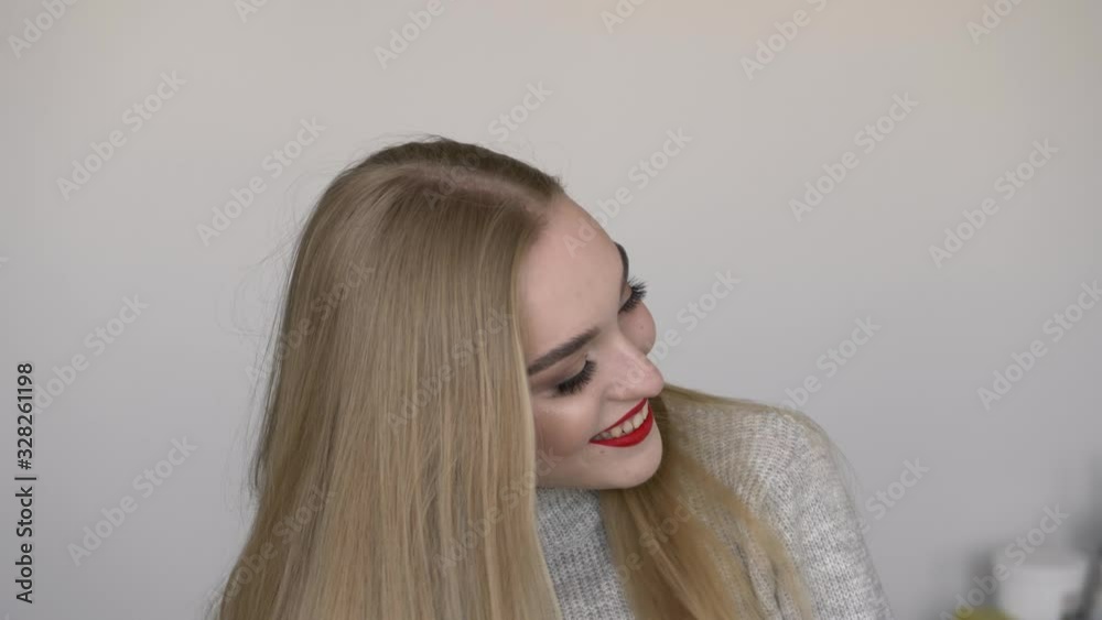 Indoors slow motion close up face shot of young smiling woman smoothing her long blond hair by hand, bright red lips, dressed grey sweater, posing against white wall natural light, model looking into Indoors slow motion close up face shot of young smiling woman smoothing her long blond hair by hand, bright red lips, dressed grey sweater, posing against white wall natural light, model looking into
