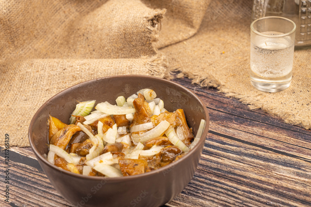 Salted mushrooms with onions in a ceramic bowl , a glass of vodka and a decanter on a background of rough homespun fabric. Close up. Salted mushrooms with onions in a ceramic bowl , a glass of vodka and a decanter on a background of rough homespun fabric. Close up.