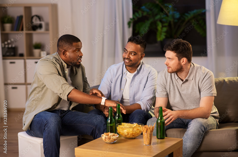 friendship, leisure and people concept - happy male friends with smart watch drinking beer and talking at home at night