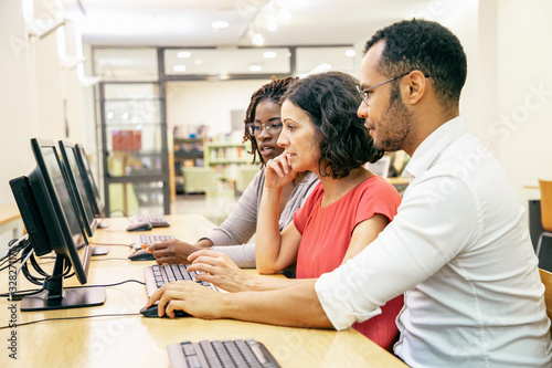 Diverse adult college mates working together in computer class. Man and women in casual sitting at table, using desktop and discussing content on monitor. Students teamwork concept