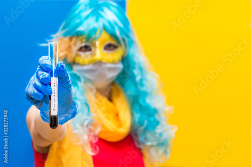 Fototapeta Naklejka Na Ścianę i Meble -  Concept of protection against coronavirus at the carnival venice. A bright woman in a blue wig, yellow mask and medical gloves holds a test tube with a blood test for Virus crown. Copy space