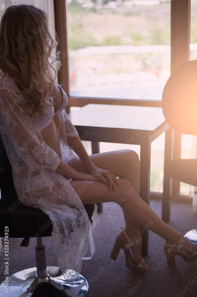 Beautiful young woman in white lace peignoir and lingerie sits in chair near window in the bedroom Beautiful young woman in white lace peignoir and lingerie sits in chair near window in the bedroom