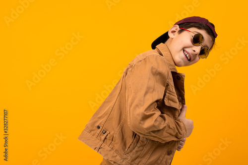 stylish teenager boy in a brown denim jacket and stylish glasses smiles and stands sideways on a yellow background with copy space © Ivan Traimak