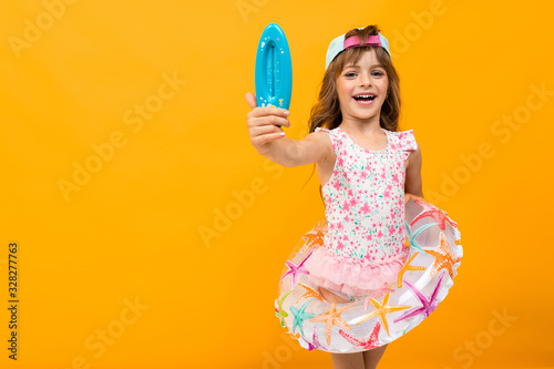 Beautiful little girl in swinsuit holds a rubber ring and shows a thermometer for a water isolated on yellow and orange background photo