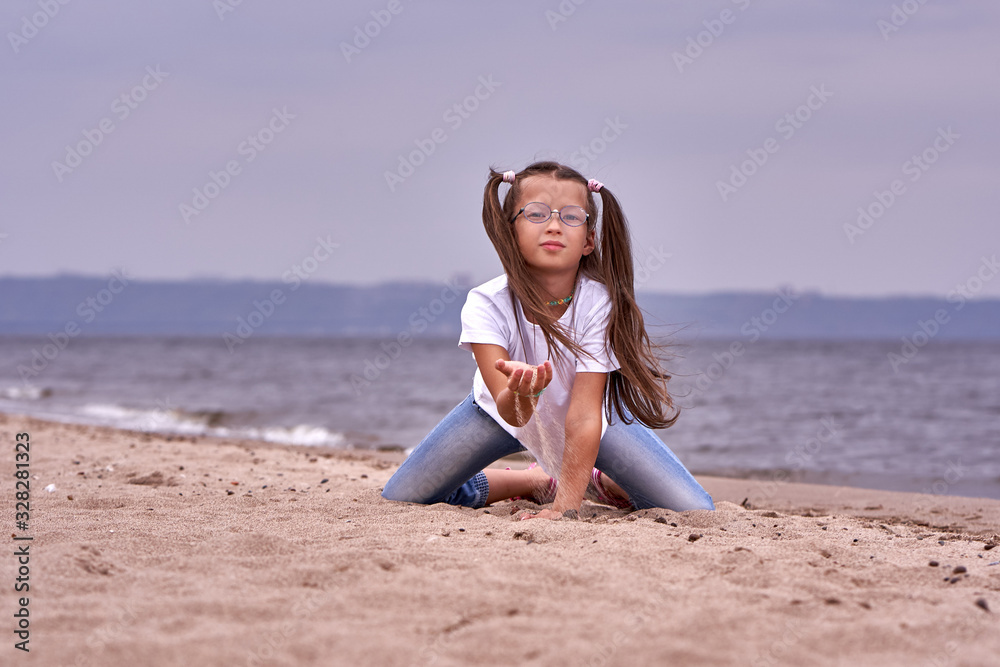 Portrait of a teenage girl in glasses and a white T-shirt walking on a sandy shore on a cloudy autumn day.