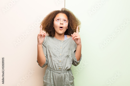 african american little girl feeling awed and open mouthed pointing upwards with a shocked and surprised look against flat wall photo