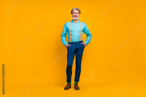 Full length photo of cool trendy grandpa hold hands pockets charismatic person stylish look clothes wear shirt suspenders bow tie pants boots socks isolated yellow color background