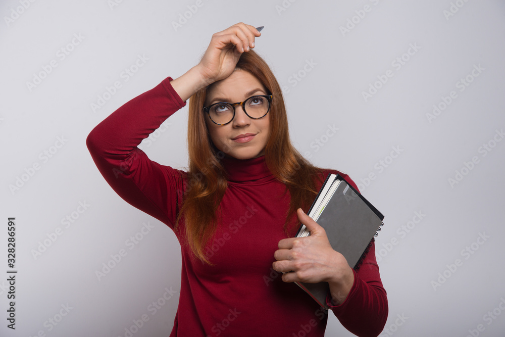 Naklejka premium Thoughtful female student with textbooks and pen. Beautiful tired young woman in eyeglasses holding notebooks and looking up isolated on white background. Education concept