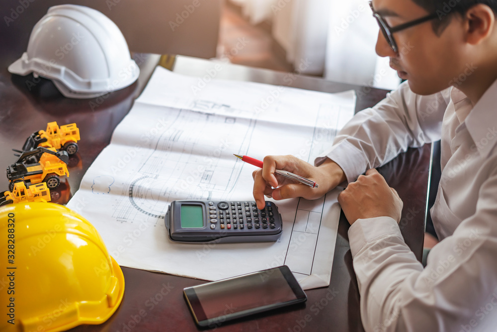 Φωτογραφία Architecture man using a calculator and pressing on the buttons with a pen in his hand and blueprint of an architectural design, sitting on a table with smartphone and yellow hardhat in foreground