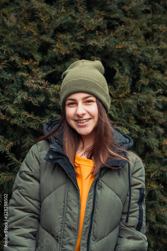 Vertical Portrait of a beautiful smiling girl with braces in a yellow sweater and khaki hat who stands near a Christmas tree in the fresh air