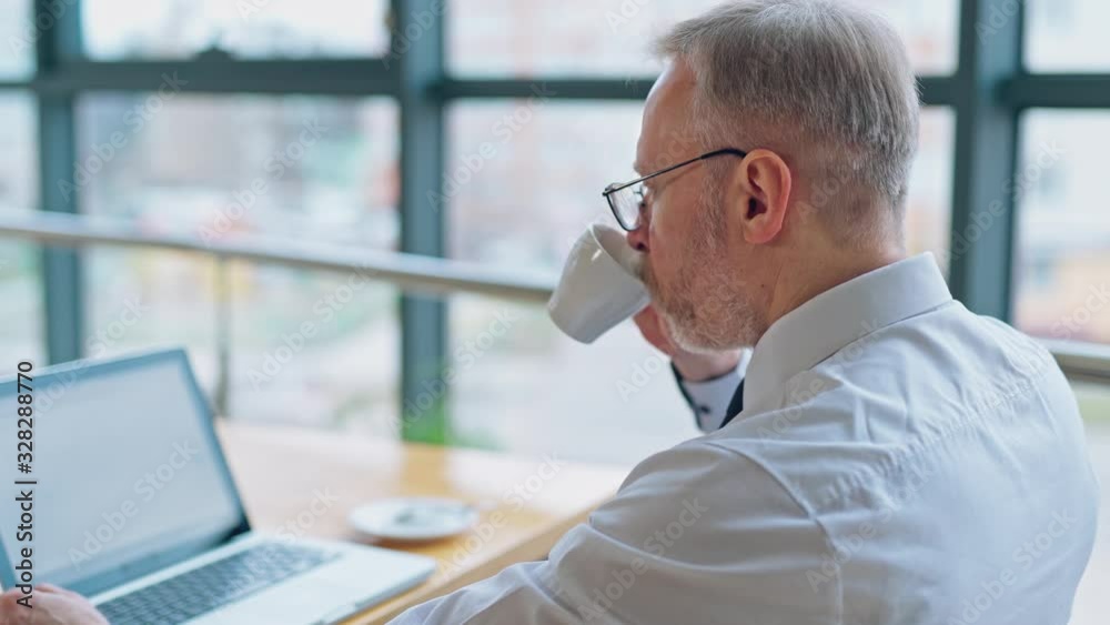 Businessman is checking presentation on a laptop while sitting at a table in light room with panoramic business view. Business video. Businessman is checking presentation on a laptop while sitting at a table in light room with panoramic business view. Business video.
