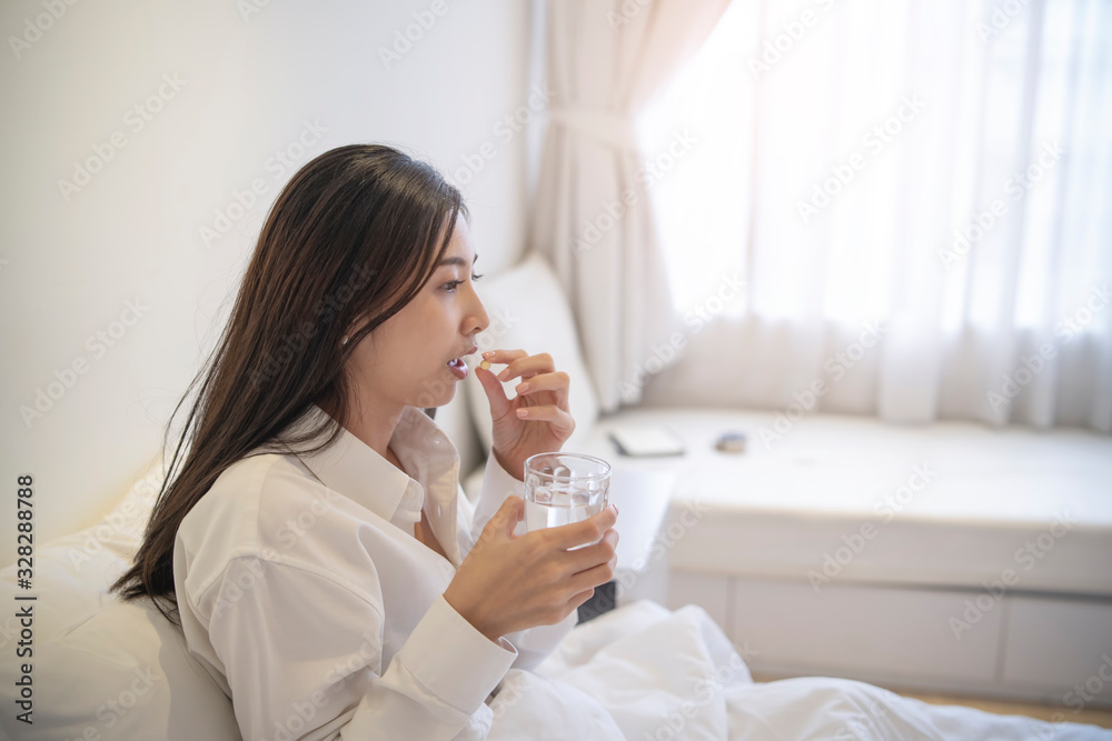 asian woman feeling sick putting a pill into mouth and holding a glass of drinking water lying in the bed be covered with blanket over, leaning on a pillow in the bed with lights through the window