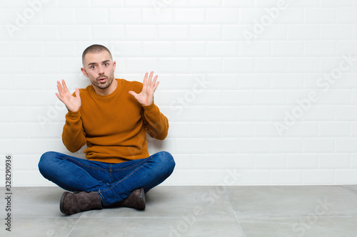 young handsome man looking shocked and astonished, with jaw dropped in surprise when realizing something unbelievable sitting on the floor photo