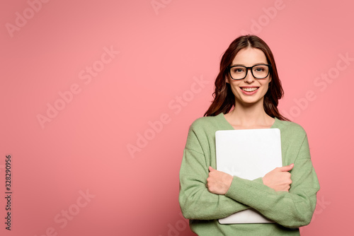 happy businesswoman in glasses holding closed laptop while smiling at camera on pink background