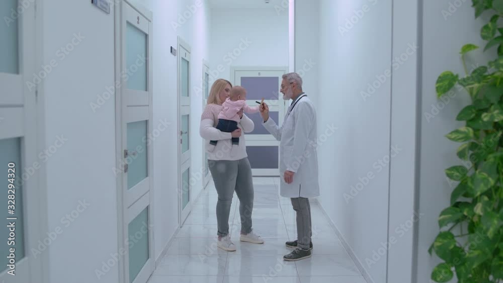 Cute active baby in mother arms playing with friendly senior doctor in clinic Cute active baby in mother arms playing with friendly senior doctor in clinic
