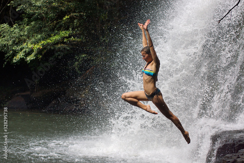 Young athletic woman jumping into water with raised hands. Background of many drops of waterfall and tropical forest © Vladimir