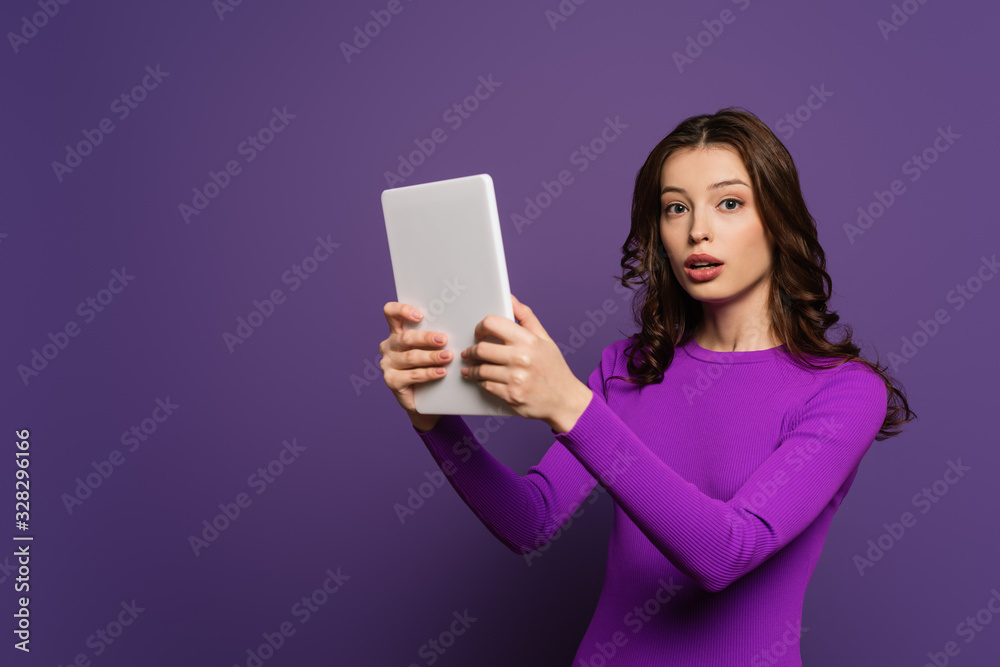 shocked girl looking at camera while holding digital tablet on purple background shocked girl looking at camera while holding digital tablet on purple background