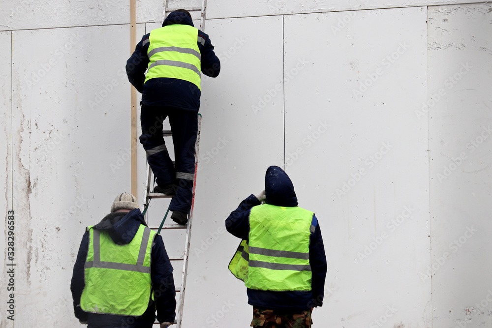 Fototapeta premium Workers near the wall of a building under construction. Man in uniform climbs the ladder