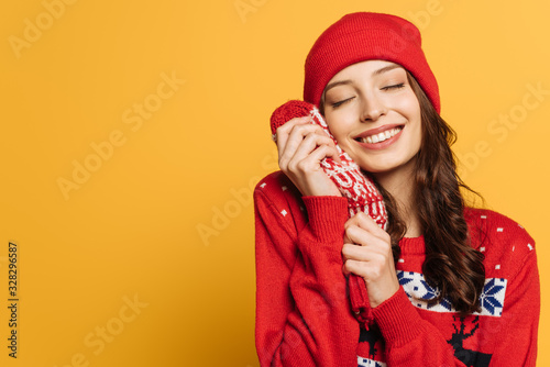 happy girl in hat and red ornamental sweater holding mittens near face with closed eyes on yellow background