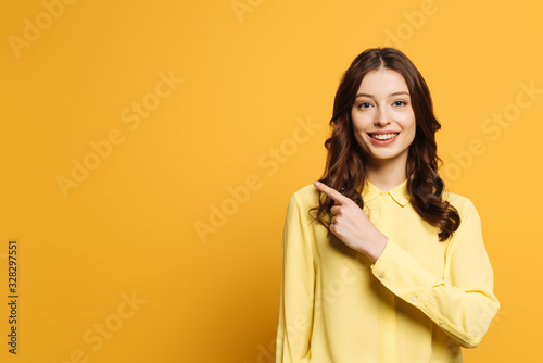 smiling girl pointing with finger while looking at camera on yellow background