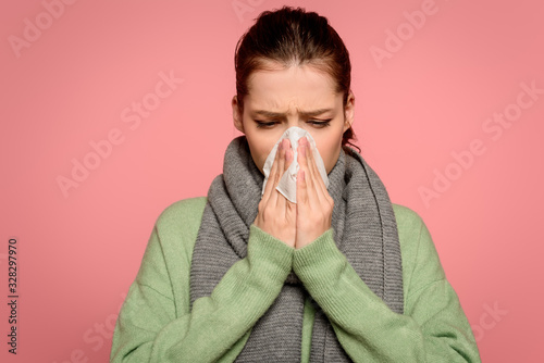sick girl in warm scarf sneezing in paper napkin isolated on pink
