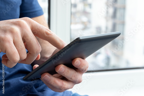 Gray e-reader electronic book in male hands. A man in a blue t-shirt with a tablet against a blurred background. Selective focus