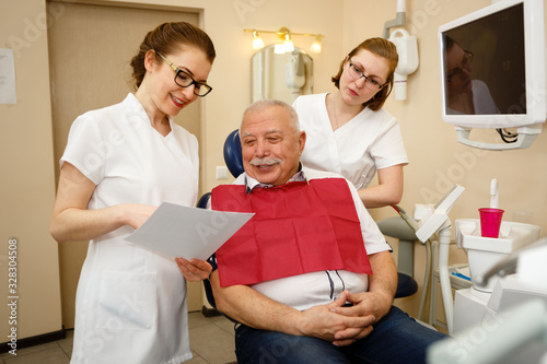 Female dentist in dental office talking with senior man 75 years old in dental armchair with napkin over his chest who is happy and pleased. Dentistry medicine and healthcare stomatology concept